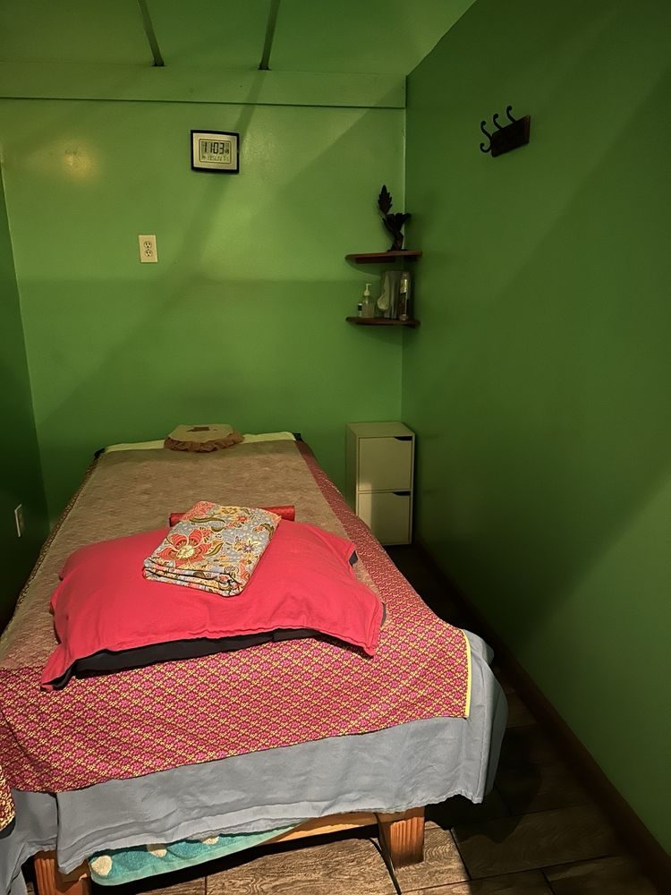 A treatment room with a massage table made up in red cloth, a folded towel at the head and a small shelf on the green wall.