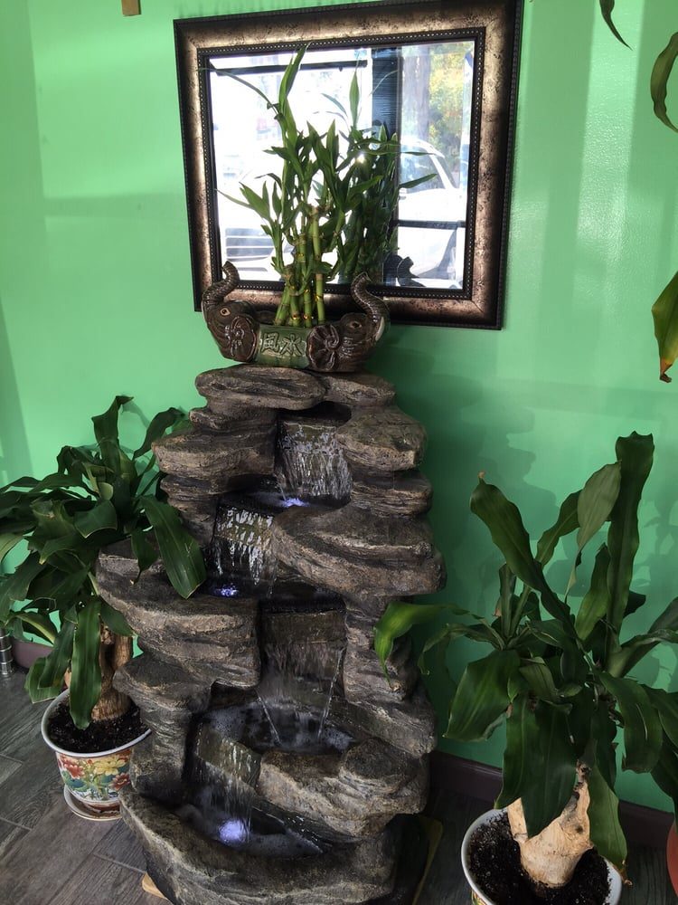 A stacked rock fountain with water running down it, potted plants on either side and a mirror above it on the green wall.