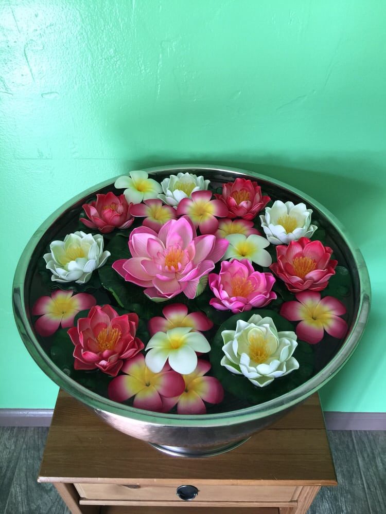 A metal bowl of pink and white lotus and plumeria flowers floating in water, on a wooden stand against the green wall.